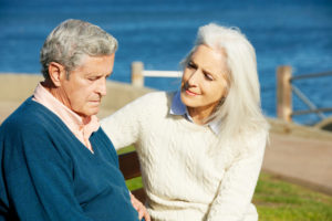 Two senior citizens sit on a bench by the sea side. One person is a senior man who looks sad. The second person is a older woman who is comforting him. 