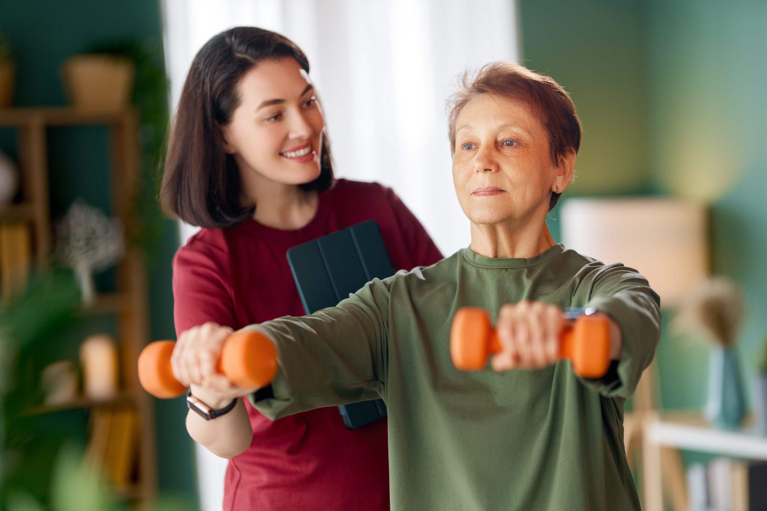 Senior woman doing dumbbell exercise while a nurse helps her with physical therapy at home.