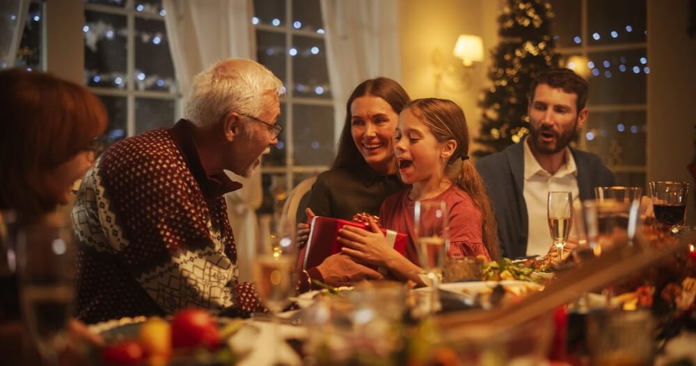 A grandfather accepts a wrapped red and white gift from his granddaughter, while her parents watch. The family is sitting at the dinner table, surrounded in food, and festive holiday decor.