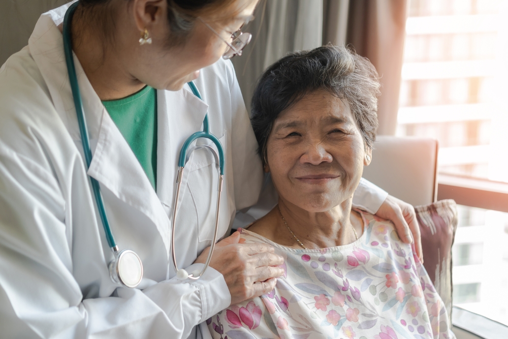 A senior woman sits and smiles at the camera while her nurse watches over her.