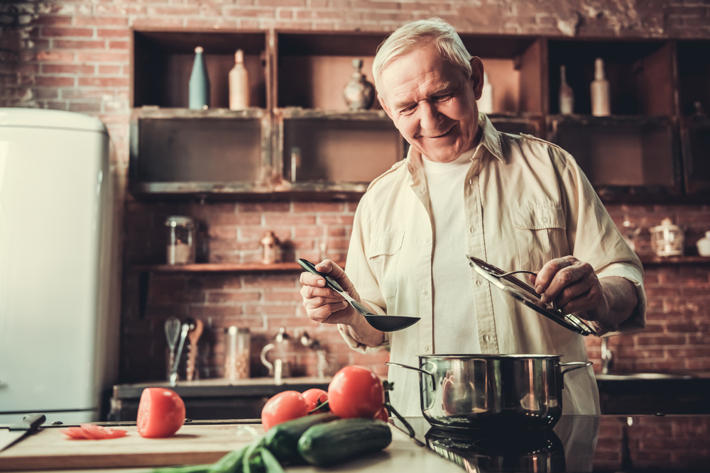 An elderly man gladly cooks on his stove with fresh produce nearby to complete the recipe.