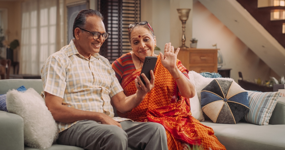 Two senior parents of Indian decent sitting on the couch talking to their relatives on the phone.