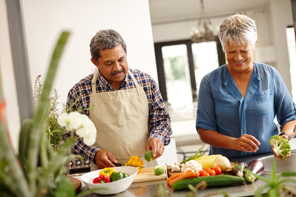 An elderly man and woman prepare vegetables on their kitchen counter by rinsing and cutting them up. They are both smiling.
