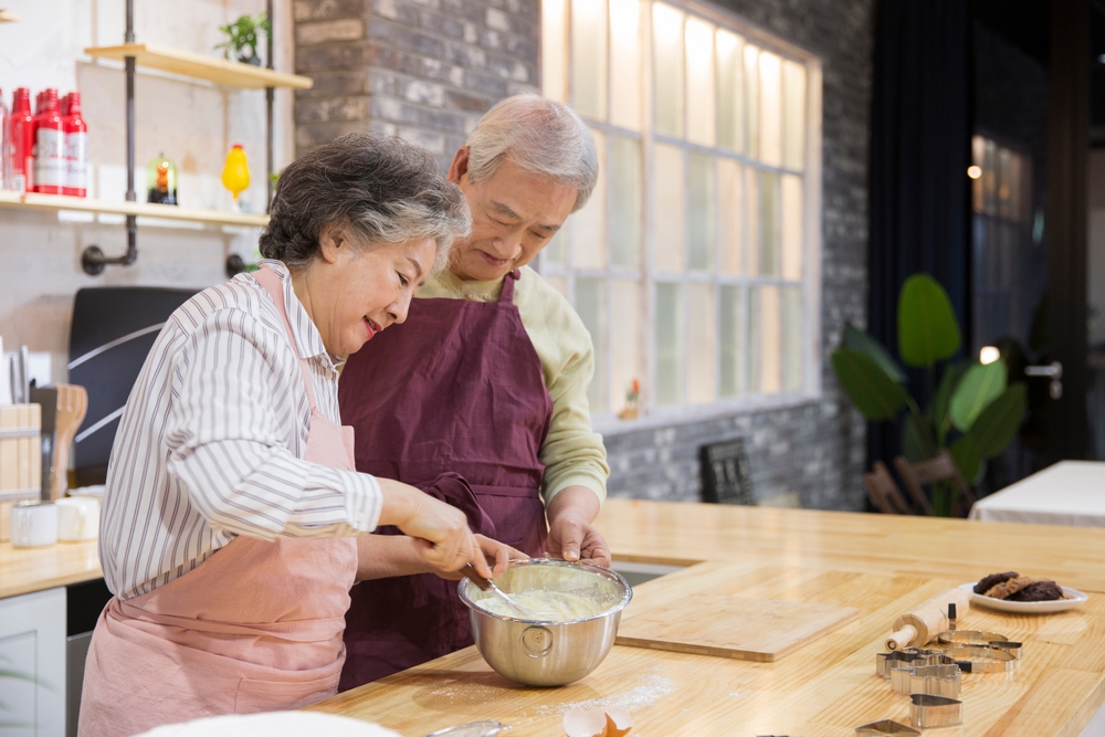 A senior couple stands in their kitchen. The wife is stirring batter in a metal bowl while the husband observes. They are both smiling.