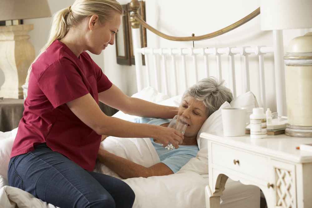 A nurse with blonde hair pulled back into a ponytail sits on the edge of the bed as she assists an elderly, bed-bound woman with a drink of water.