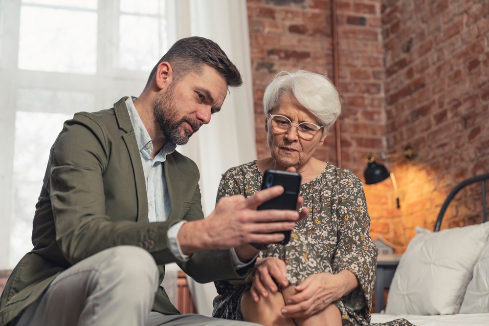 An adult child sits on the couch with his senior mother as he helps her review the apps on her phone.