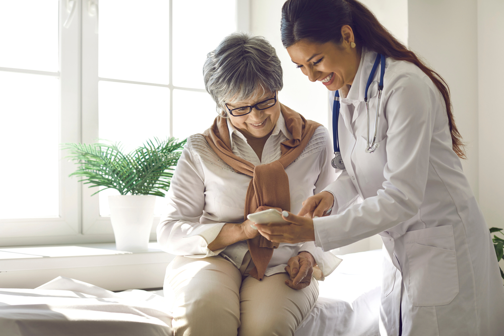 Un médecin se tient à côté d’une patiente âgée, un téléphone à la main, montrant à sa patiente comment accéder aux applications de soins de santé sur son téléphone. 