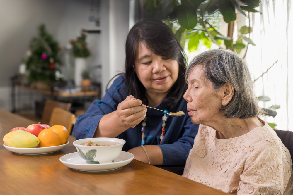 An adult sitting at a table beside an elderly woman helping her eat soup.