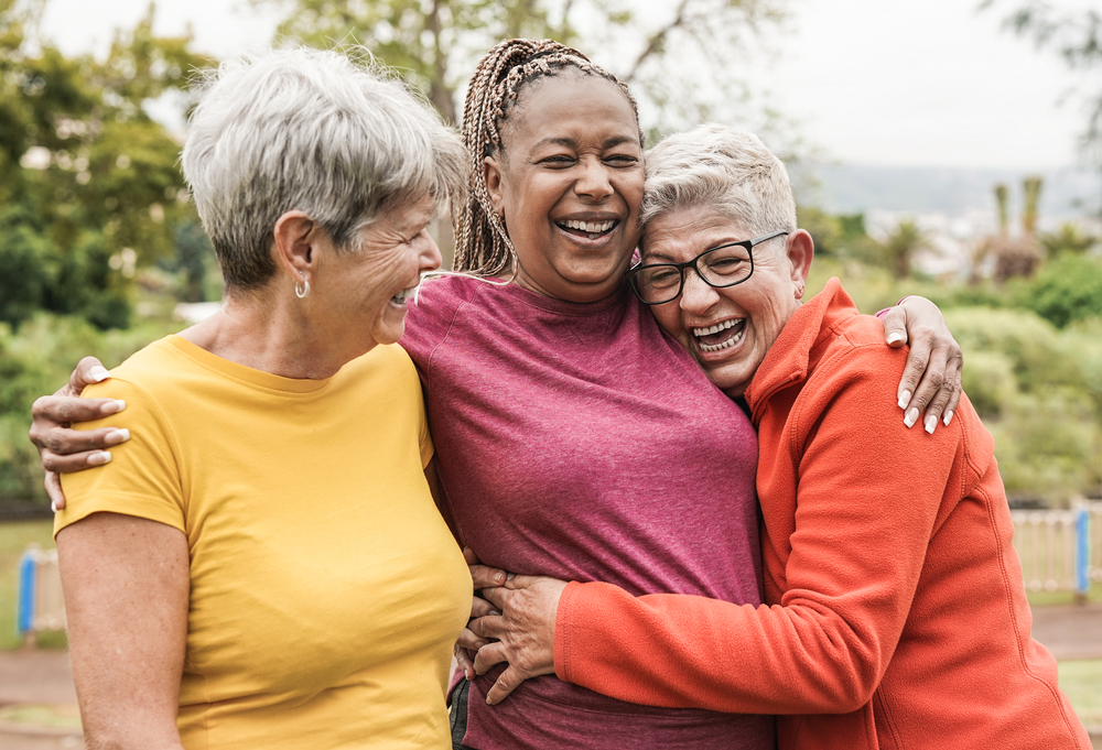 Three older senior ladies outside hugging, smiling, and enjoying life.