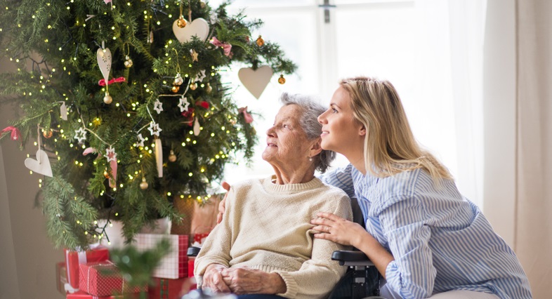 Caregiver sits beside senior during the holidays in front of a Christmas tree.