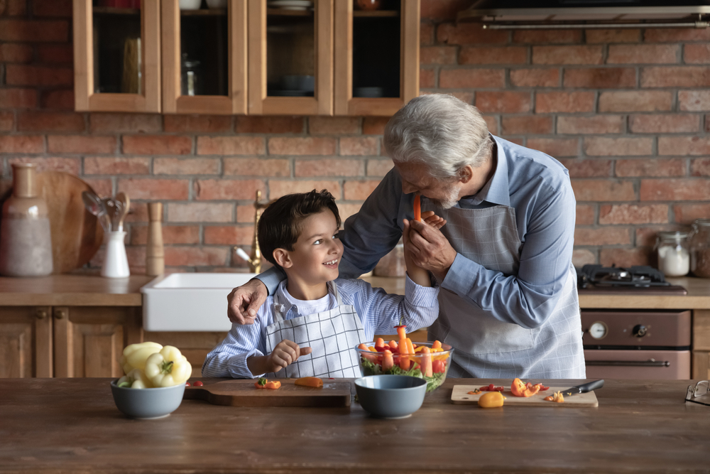 A grandfather stands in the kitchen with his grandson making a fruit salad together.