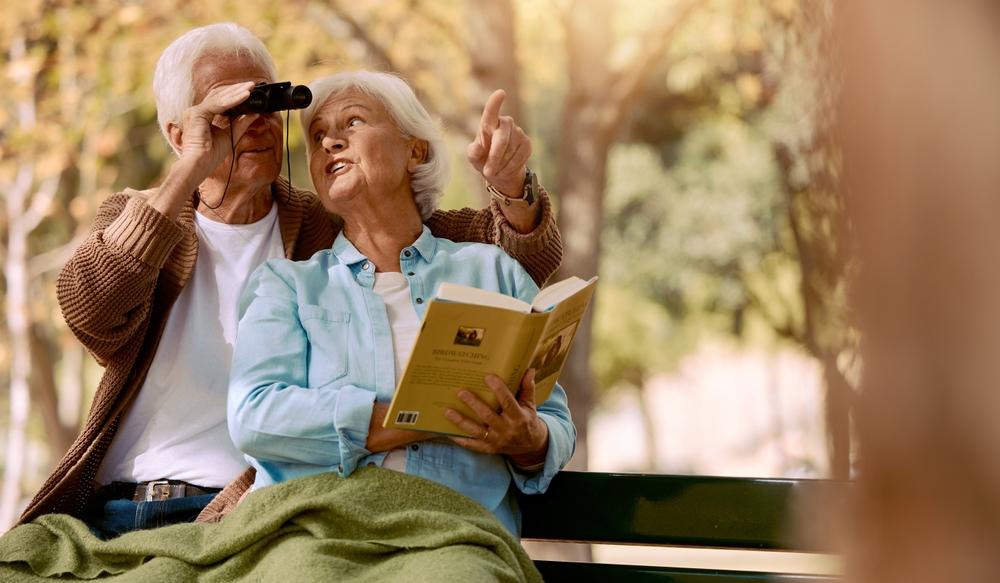 A senior couple sits outside with binoculars watching birds.